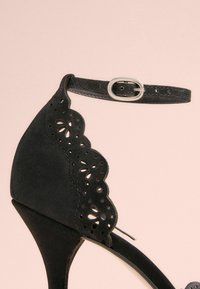Mustat korkokengät, joissa on aallotettu leikattu muotoilu ja reikiin, varustettu soljilla nilkkahihnalla ja hoikalla stiletto-kannan.