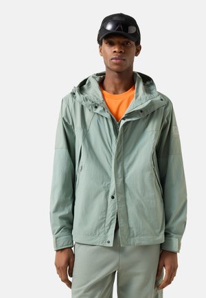 Man wearing light green hooded jacket, matching pants, orange shirt, black cap, and sunglasses on cap against light background.