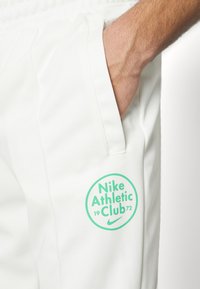 Vita träningsbyxor med elastiskt midjeband. Har en grön cirkulär logotyp med "Nike Athletic Club 1972" på låret. Slät textur.