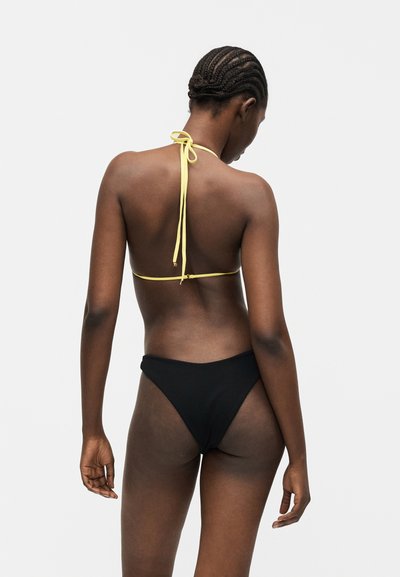 Femeie cu părul împletit, purtând un top de bikini galben halter și chiloți negri, văzută din spate pe un fundal simplu.