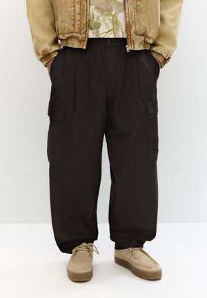 Crne cargo hlače s bočnim džepovima, opuštenog kroja i elastičnom futrom. Uparene s bež slip-on cipelama i kašmir bomber jaknom.