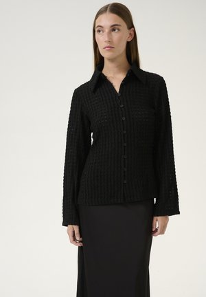 Chemise noire texturée à boutons, dotée d'un col, de manches longues et d'un motif côtelé. Portée par-dessus une longue jupe noire.