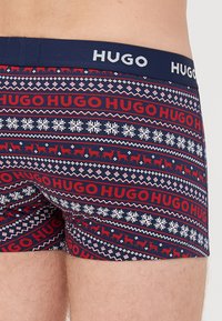 Marineblaue Boxershorts mit einem sich wiederholenden Muster aus rotem Text, Schneeflocken und weißen Rentieren. Elastischer Bund mit "HUGO"-Markenlogo.
