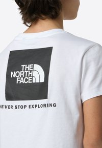 Tricou alb din bumbac, cu un grafic pătrat mare negru, pe care este scris „THE NORTH FACE” și textul „NEVER STOP EXPLORING” dedesubt, cu litere negre îndrăznețe.