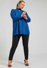 Blusa blu a maniche lunghe con scollatura a chiave e maniche arricciate, abbinata a pantaloni in ecopelle nera e tacchi neri con cinturini.