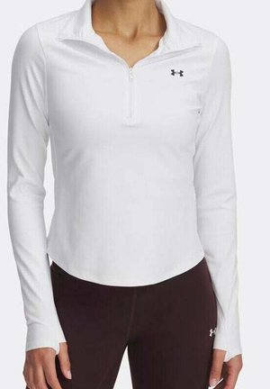 Kobieta nosząca biały sportowy top z długim rękawem, z suwakiem do połowy oraz czarnym logo Under Armour na piersi.