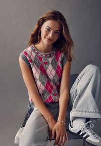 Knit sleeveless sweater featuring a pink argyle pattern with gray, accented by a round neckline with heart details; paired with gray trousers.
