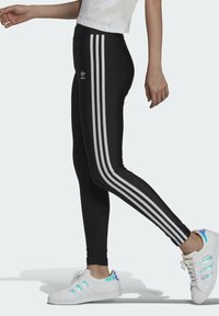Leggings negre cu trei dungi verticale albe pe lateral. Textură netedă cu un design ajustat, purtate cu adidași albi cu accente iridescente.