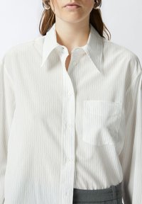 Camicia bianca a maniche lunghe con sottili righe verticali, colletto classico, chiusura frontale con bottoni e una singola tasca sul petto. Tessuto dalla consistenza liscia.