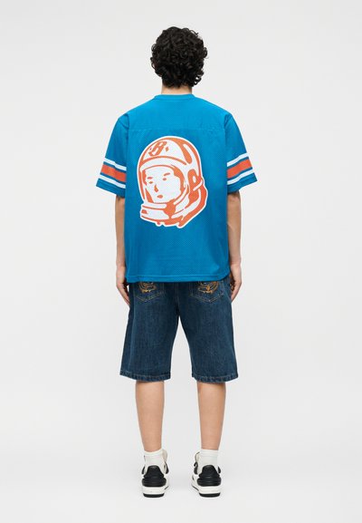 Person med krøllete hår står, iført blå kortermet trøye med oransje og hvitt astronaut-hjelmtrykk, denimshorts og joggesko.