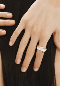 Anillo dorado y blanco con diseño entrelazado; presenta cinco pequeñas piedras integradas. La mano descansa sobre el cabello negro largo y liso.