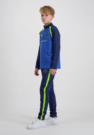 Jongen in een blauw en marineblauw trainingspak met neon groene strepen en witte sneakers, staand zijwaarts tegen een effen achtergrond.