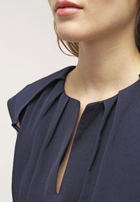 Tmavě modrá halenka s plisovaným výstřihem, krátkými rukávy a detailním průstřihem. Látka má hladkou texturu a strukturovaný design.