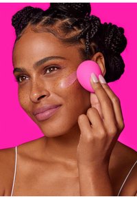 Una spugna per il trucco rosa tenuta da una persona che applica il prodotto sulla guancia. Lo sfondo è di un rosa brillante; la pelle ha una texture naturale e liscia con lentiggini.