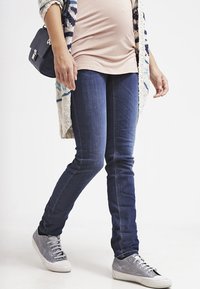 Jeans skinny azul escuro com uma textura suave, combinados com sapatilhas cinzentas. O conjunto apresenta uma blusa rosa clara e um cardigan estampado.