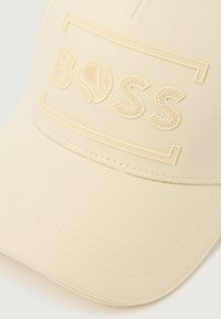 Bež baseball kapa s teksturiranim logotipom "BOSS" i grafikom teniskog lopta urezanom na prednjem panelu.