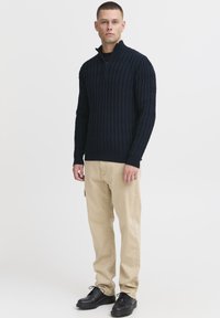Suéter de punto azul marino con cuello de media cremallera, que exhibe un patrón texturizado. Combinado con pantalones beiges y zapatos negros de cordones.