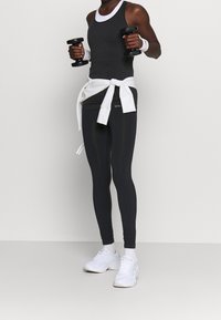 Svart atletisk linne med vit krage, i kombination med höga svarta leggings och vita sneakers. Håller hantlar, knyter en vit sweatshirt i midjan.