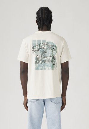 Homme aux cheveux tressés portant un t-shirt blanc avec un motif bleu et le texte « Stillness in Motion », associé à un jean bleu clair, de dos.