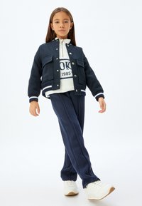 Giacca blu scuro con due tasche frontali e bottoni argentati, abbinata a una felpa bianca con logo navy e pantaloni blu navy abbinati e scarpe bianche.