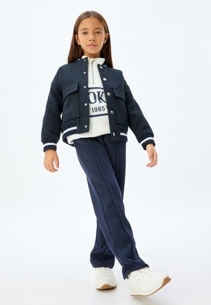 Giacca blu scuro con due tasche frontali e bottoni argentati, abbinata a una felpa bianca con logo navy e pantaloni blu navy abbinati e scarpe bianche.