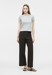 Jeune femme debout, portant un t-shirt gris clair à manches courtes, un pantalon noir à jambes larges avec poches avant, et des tongs noires.