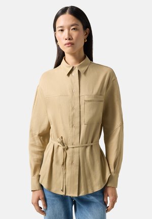 Kobieta ubrana w beżową koszulę na guziki z przednią kieszenią i paskiem do wiązania, w połączeniu z niebieskimi dżinsami, stojąca na jednolitym tle.