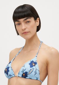Blå blommig bikiniöverdel med triangeldesign och justerbara remmar, prydd med mörkblå blommor mot en ljusblå bakgrund.