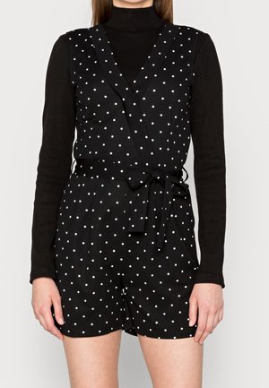 Femme portant un col roulé noir à manches longues sous une combinaison sans manches noire à pois avec une ceinture nouée à la taille, debout devant un fond clair.