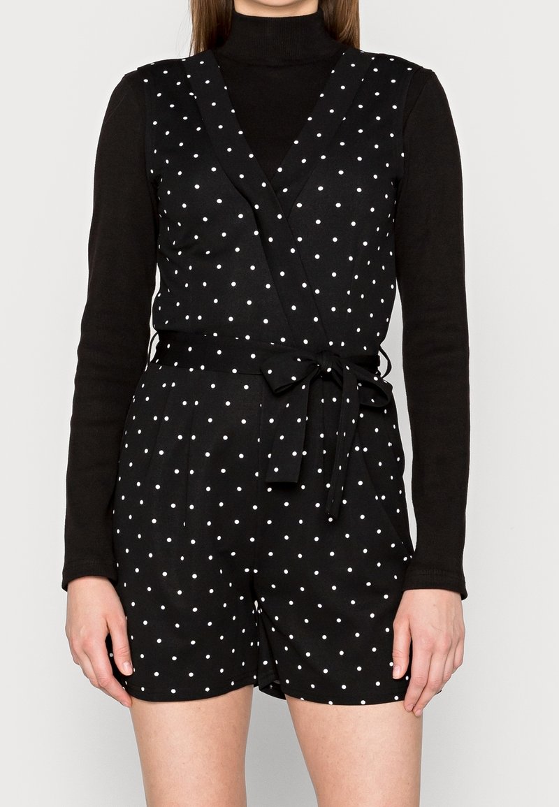 Femme portant un col roulé noir à manches longues sous une combinaison sans manches noire à pois avec une ceinture nouée à la taille, debout devant un fond clair.