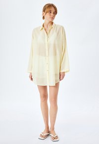 Camicia gialla a maniche lunghe, con bottoni, realizzata in tessuto trasparente, con una vestibilità rilassata e una leggera lucentezza, abbinata a ciabatte bianche.