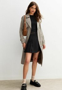 Lange geruite trenchcoat over een zwarte cropped top en een zwarte geplooide rok. Zwarte loafers met gouden details. Bruine schoudertas en beige sokken.