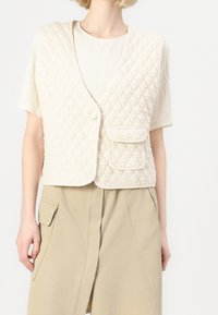 Chaleco acolchado crema con escote en V, que presenta un cierre de botones y un bolsillo delantero, llevado sobre una camisa blanca texturizada y pantalones beiges.