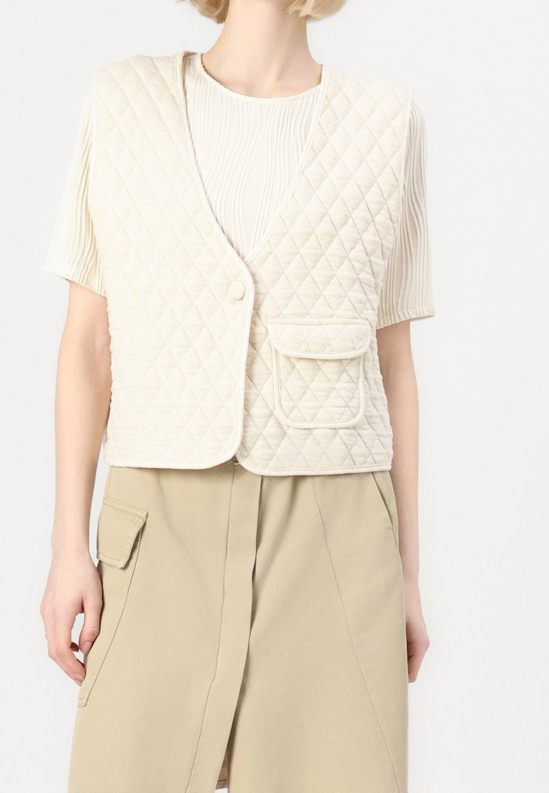 Chaleco acolchado crema con escote en V, que presenta un cierre de botones y un bolsillo delantero, llevado sobre una camisa blanca texturizada y pantalones beiges.