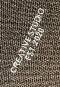 Λευκό κεντημένο κείμενο "CREATIVITY STUDIO EST 2020" σε υφασμάτινο, καφέ φόντο με υφή, τοποθετημένο διαγώνια.