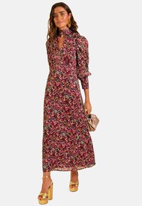 Vestido largo con estampado floral, mangas abullonadas, escote en V profundo y lazada en el cuello alto. Los colores incluyen burdeos, rosa y verde. Tacones de plataforma dorados.