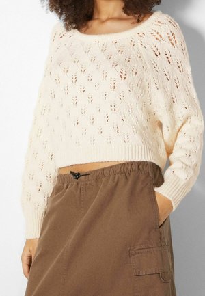 Frau trägt einen cremefarbenen, gestrickten, kurz geschnittenen Pullover mit Blattmuster und einen hoch taillierten braunen Rock mit Taschen, Hand in der Tasche.