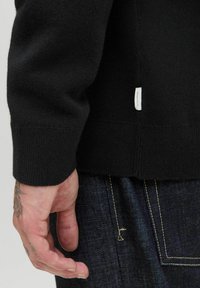 Black knit sweater with a ribbed cuff, textured fabric, and a small white brand label on the side. Dark denim pants are partially visible.