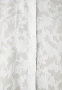 White sheer blouse featuring a textured floral pattern and button closure. Soft fabric with subtle layers and a lightweight appearance.