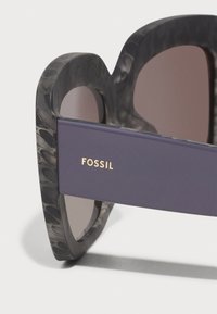 Fossil solbriller med en grå mønstret stel og lilla accent. Linserne er farvet brune, hvilket fremhæver en glat tekstur og afrundet design.