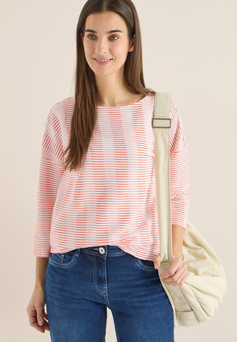 Smilende kvinde med langt brunt hår iført en rød- og hvidstribet top og blå jeans, bærende en beige skuldertaske på højre side.