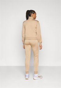 Beige träningsdräkt med hög krage och åtsittande muddar. Modellen bär vita sneakers med ljusa detaljer och tre ränder.