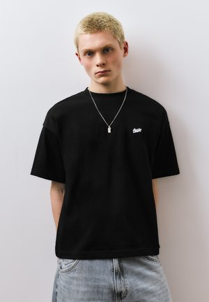 Young man with short blonde hair wears black oversized t-shirt with small white logo and silver necklace, standing against plain wall.