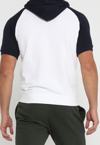 Mężczyzna w białej bluzie z krótkim rękawem i czarnymi rękawami oraz kapturem, w ciemnozielonych spodniach, widziany z tyłu na jednolitym tle.