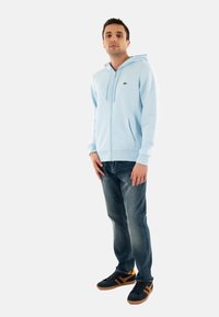 Sudadera con cremallera azul claro y bolsillo canguro, combinada con jeans oscuros y zapatillas negras con acentos marrones.