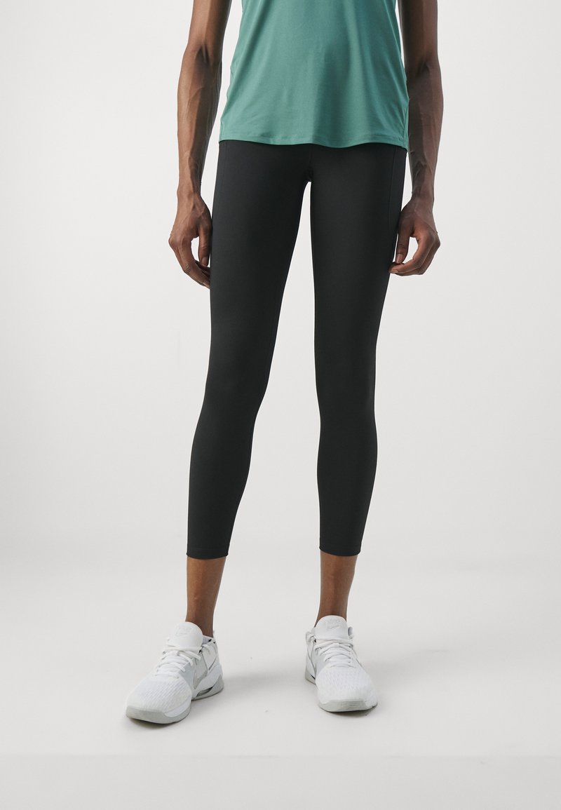Leggings neri, di lunghezza alla caviglia, design aderente, texture liscia. Abbinati a una maglietta teal e scarpe da ginnastica bianche, in piedi su uno sfondo neutro.