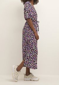 Robe fleurie avec un fond noir, des manches courtes bouffantes et une ceinture à la taille. Ornée de fleurs violettes et roses ; fentes jusqu'à la cheville.