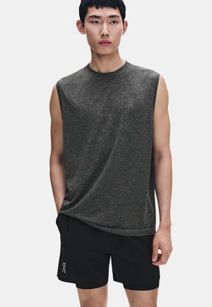 Homme portant un haut de sport sans manches gris foncé et un short noir, debout avec une main dans la poche, sur un fond clair.