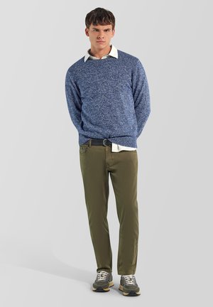 Joven de pie con las manos detrás de la espalda, vistiendo un suéter azul sobre una camisa blanca, pantalones verde oliva y zapatillas grises sobre un fondo gris.