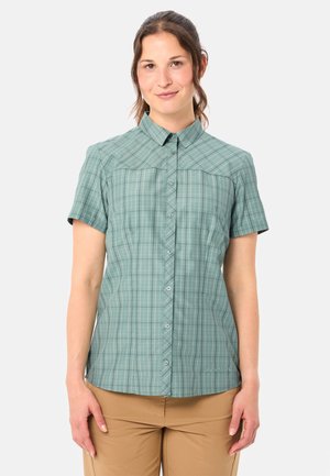Femme portant une chemise à carreaux verte à manches courtes et boutonnée, ainsi qu'un pantalon beige, debout devant un fond clair uni.